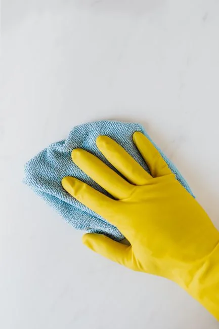 A person wipes a laptop screen using a blue cloth and spray bottle on a wooden table for hygiene.